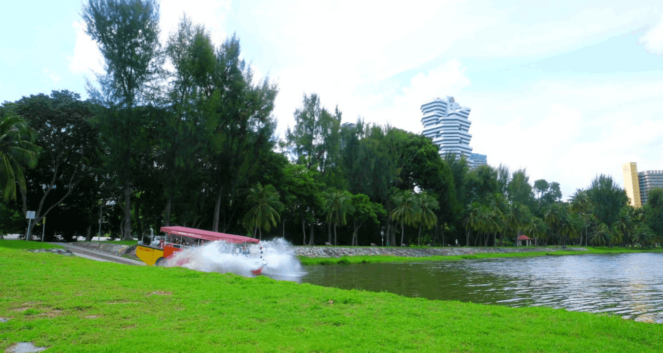 Captain Explorer DUKW Tour amphibious vehicle making a splash as it enters the water in Singapore, with lush greenery, palm trees, and modern buildings in the background.