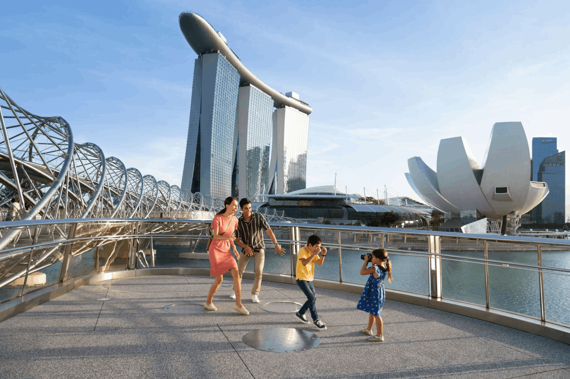 Family with children taking photos at The Helix Bridge with Marina Bay Sands and ArtScience Museum in the background.