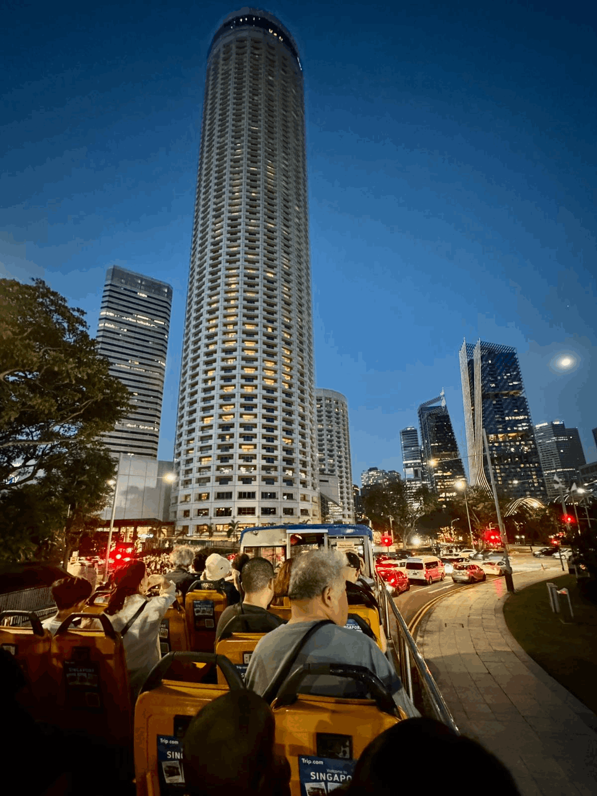 Tourists on the upper deck of the FunVee Open-Top Bus taking photos of Fairmont Singapore and Raffles City at night.