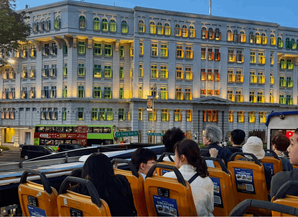 Passengers on an open-top double-decker FunVee bus viewing the colorful, illuminated windows of the Old Hill Street Police Station at dusk in Singapore.