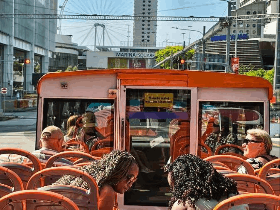 Tourists seated on the top deck of the FunVee Open Top Bus enjoying a sunny sightseeing tour in Singapore, with the Singapore Flyer and city skyline visible in the background.