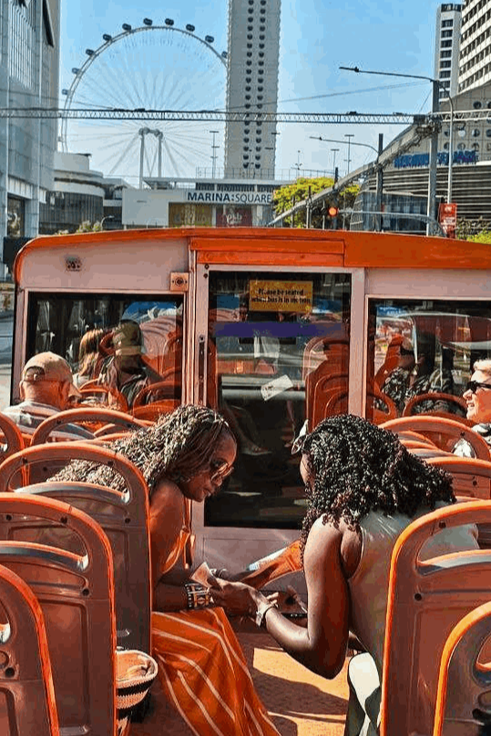 Passengers seated on the upper deck of a FunVee open-top sightseeing bus in Singapore, with a clear view of the Singapore Flyer and Marina Square in the background under a sunny blue sky.