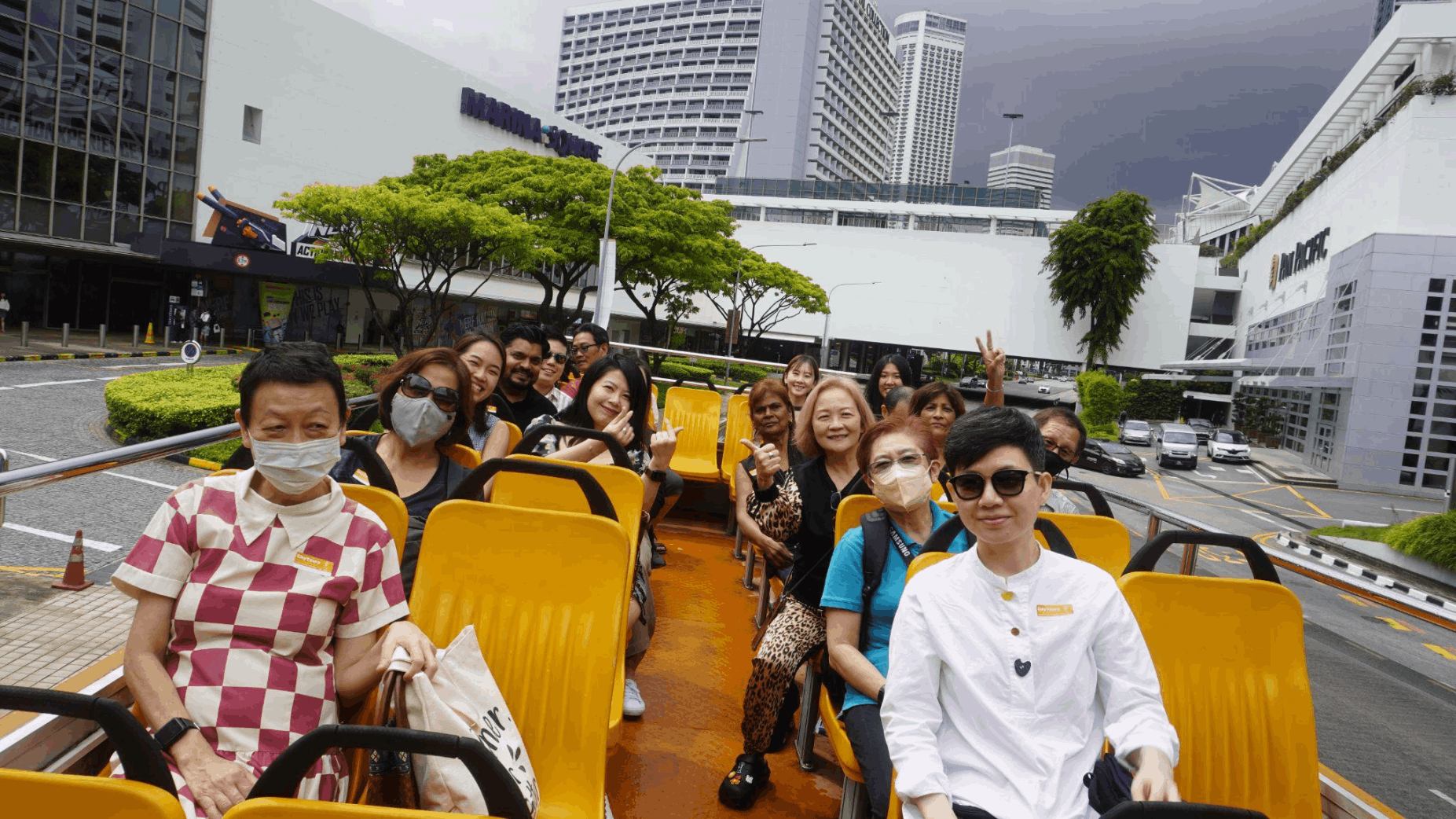 A group of people, smile and pose for a photo on the top deck of FunVee opentop bus driving through a city street in Singapore.