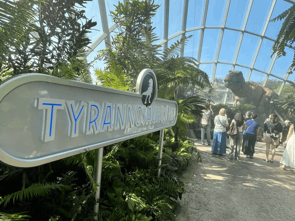 Visitors walking through the Jurassic World Experience inside the Flower Dome at Gardens by the Bay, featuring a life-sized Tyrannosaurus Rex display.