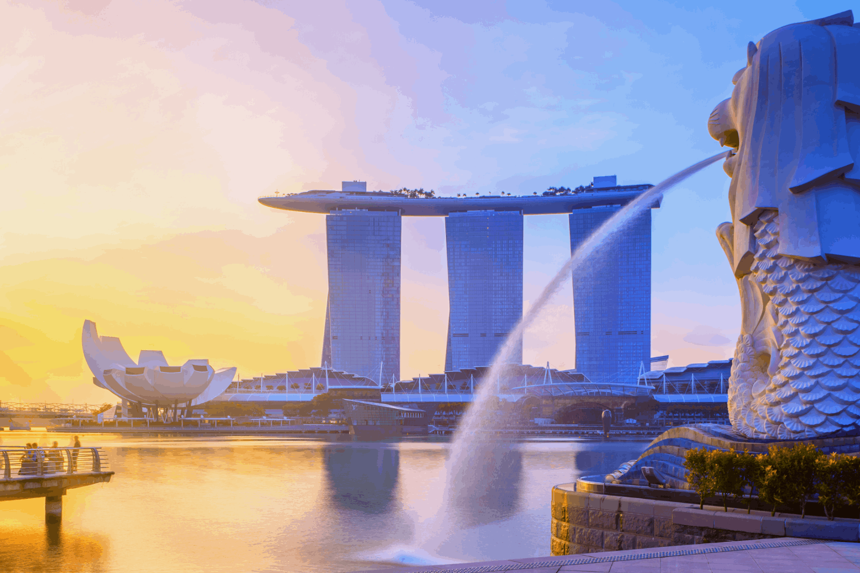 A wide-angle sunrise view of the Merlion statue spouting water at Merlion Park, with the Marina Bay Sands and ArtScience Museum reflecting on the calm water in Singapore.