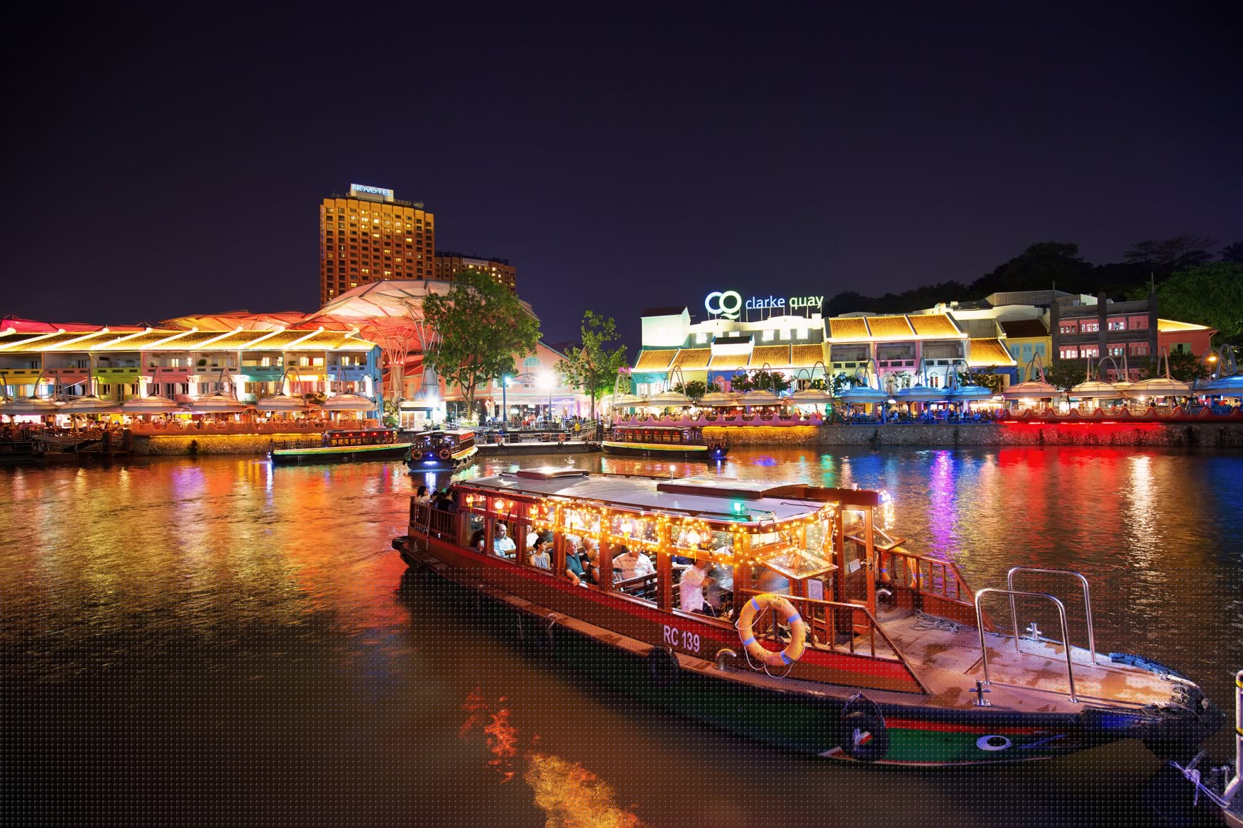 A night view of Clarke Quay in Singapore with colourful lights reflecting on the river and a traditional bumboat illuminated for the Singapore River Cruise.