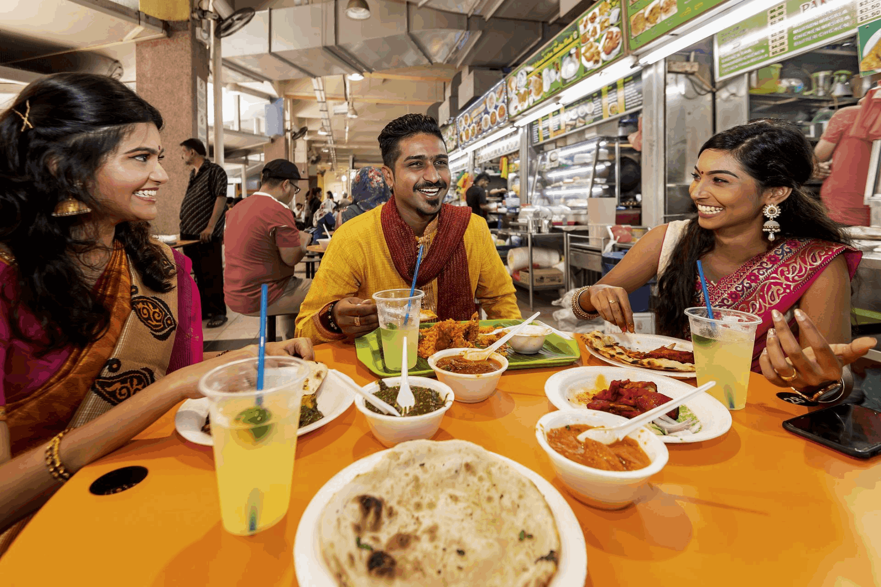 Friends enjoying Indian dishes like biryani, prata, and curry at Tekka Market in Little India, Singapore, surrounded by vibrant hawker stalls.